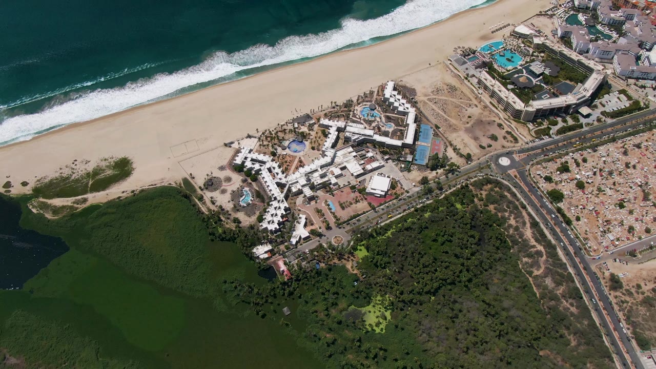 Aerial view of beach, resort buildings, road, ocean surf, and green water.