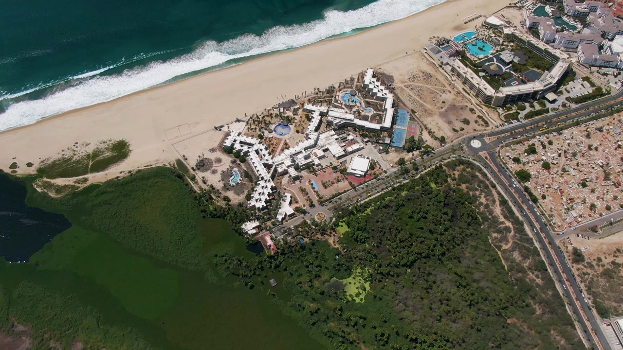 Aerial frame over San Jose del Cabo with the resort centered below the beach, white surf at the shoreline, road traffic to the right, and no visible people.