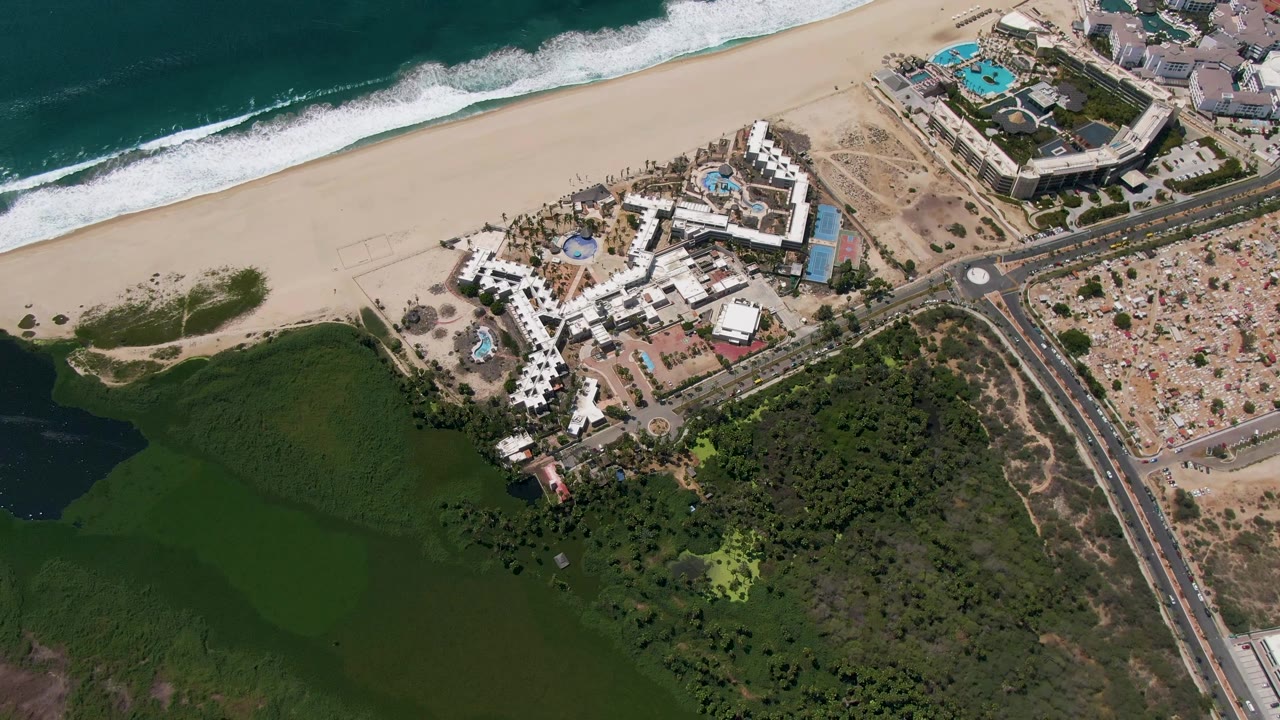 Mid-clip aerial frame in San Jose del Cabo showing ocean whitecaps along the beach, resort buildings below, the road and tan lots to the right, and no visible people.