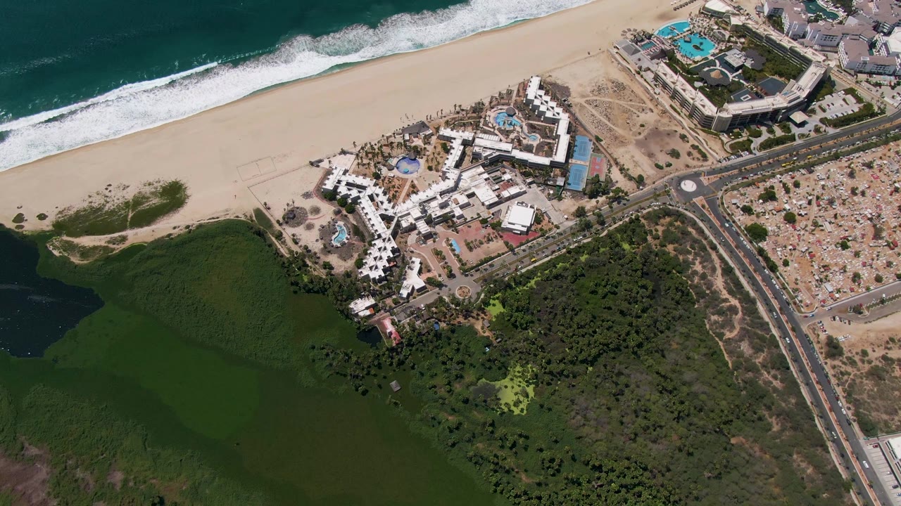 Later aerial frame over San Jose del Cabo with the beach and surf running across the top, resort buildings and road through the center, green water below, and no visible people.