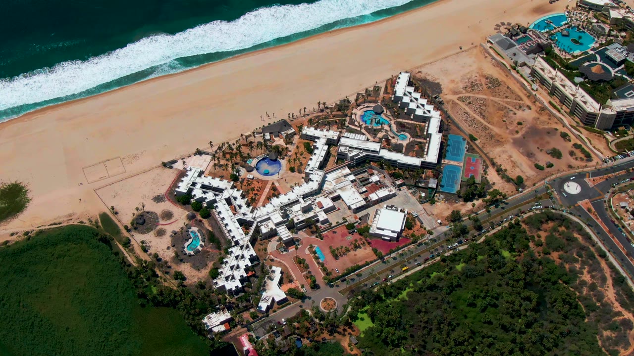 Overhead aerial frame of a San Jose Del Cabo beachfront resort with white roofs, blue pools, a sandy beach, ocean surf, roads, and greenery, with no visible people.