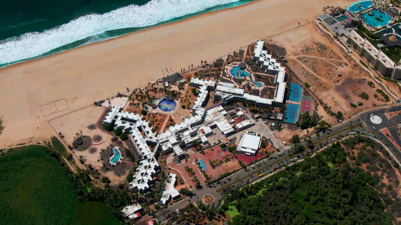 Aerial resort complex with pools, beach, road, and ocean waves.