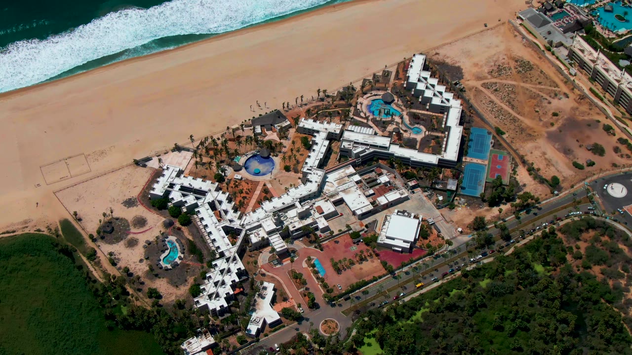 Overhead San Jose Del Cabo frame with the beach and white water across the top, resort buildings and pools in the center, road at lower right, and no visible people.