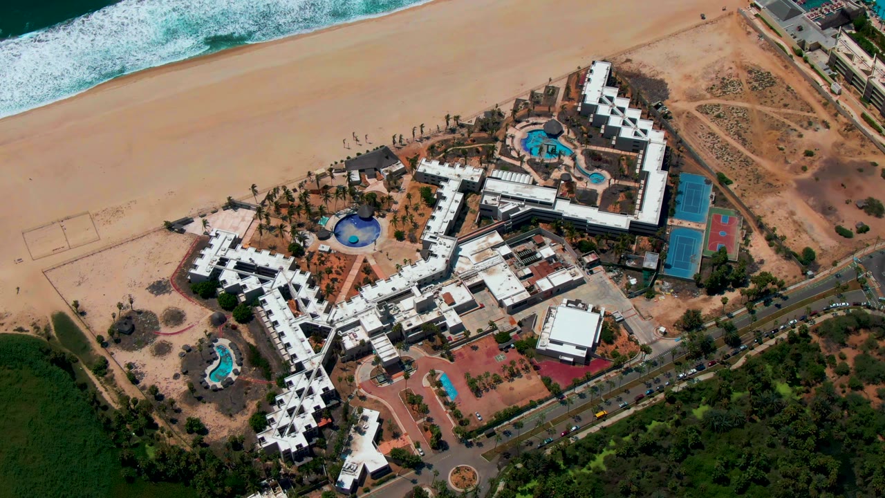 Overhead view in San Jose Del Cabo, Baja California Sur, Mexico showing white resort buildings, blue pools, sandy beach, surf, road, and green vegetation with tiny people visible.