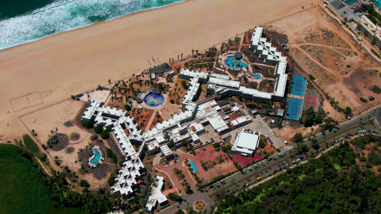 Drone view in San Jose Del Cabo with white hotel buildings and bright blue pools set between the sandy shoreline, foaming waves, a roadside edge, and green land.