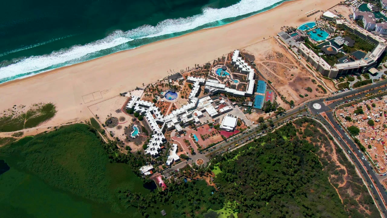 Aerial frame over San Jose del Cabo showing ocean surf, a broad sandy beach, white resort buildings with pools, a road, and green water landscape below, with no visible people.