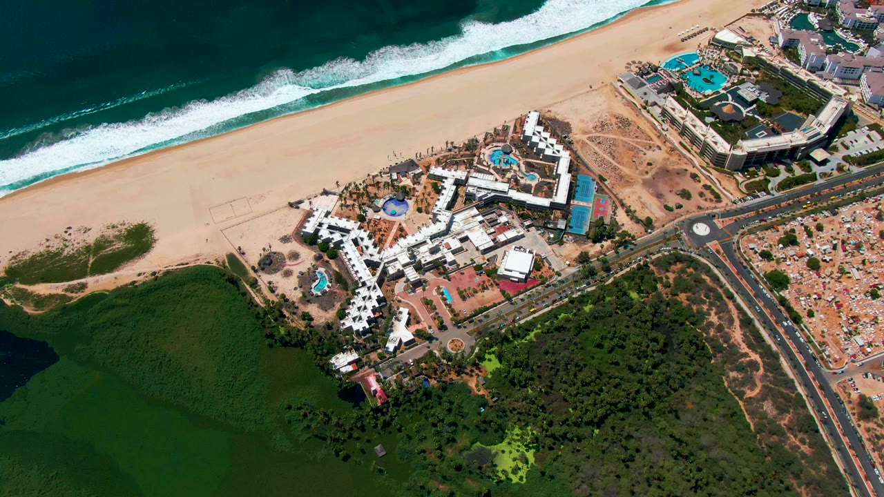 Second aerial frame in San Jose del Cabo with the resort slightly centered between the beach and road, ocean waves breaking above and green lagoon-like water below, with no visible people.