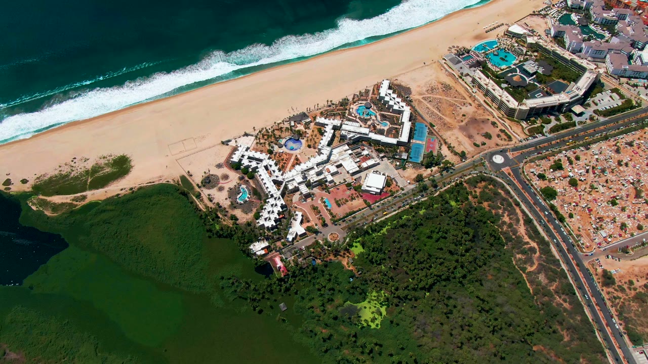 Aerial view in San Jose del Cabo with the resort near the center, a road and intersection at right, additional buildings near the beach, surf at the top, and no visible people.