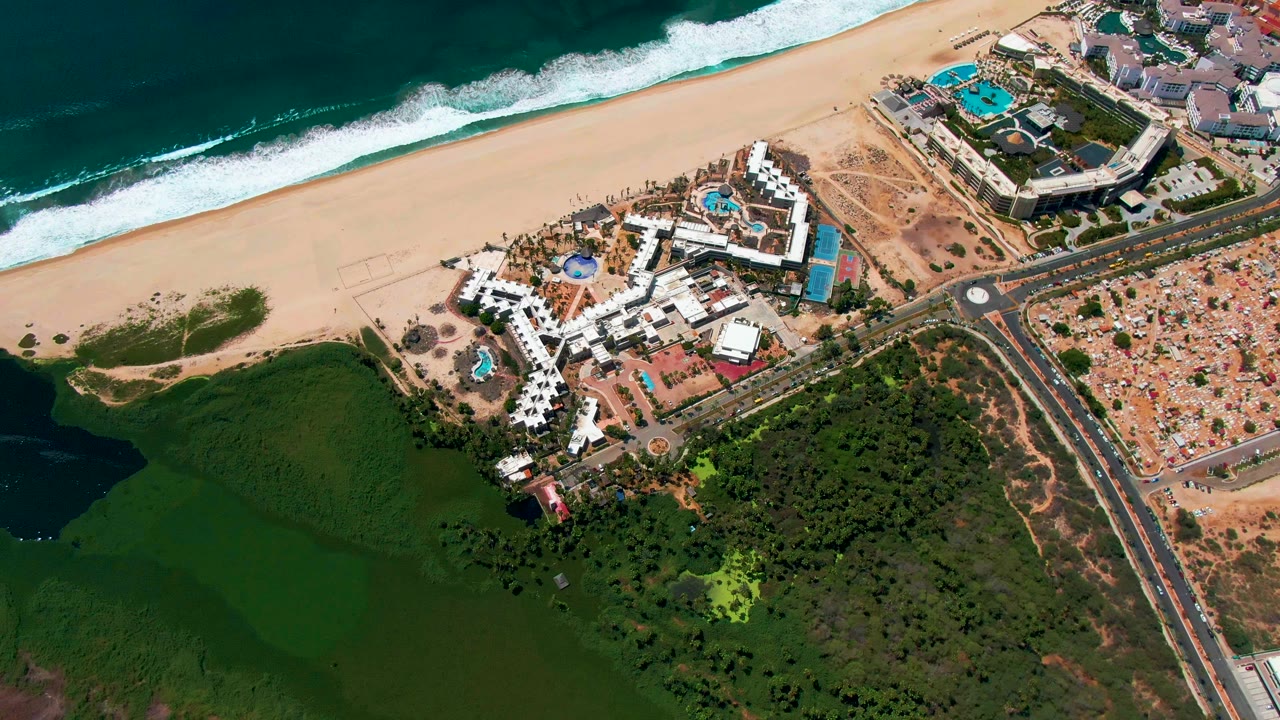 Wide aerial of resort buildings, sandy shoreline, ocean waves, road, vehicles, and green water.
