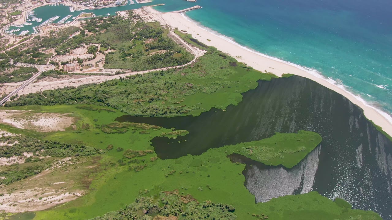 San Jose Del Cabo aerial frame showing the green cliff top over dark lagoon water, pale beach, blue-green ocean, inland roads, buildings, and no visible people.