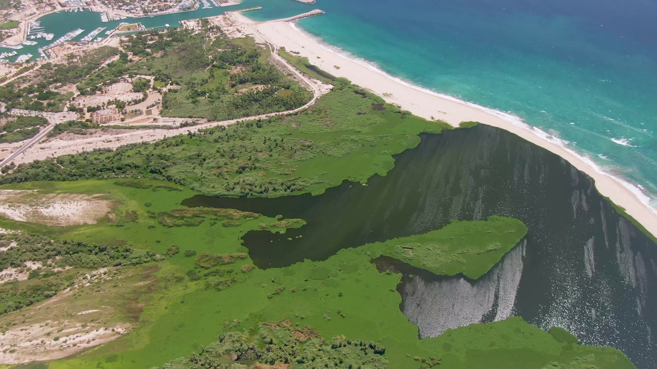 Aerial view over San Jose Del Cabo with more marshland in the foreground, a dark lagoon in the middle, white beach and turquoise ocean beyond, and no visible people.