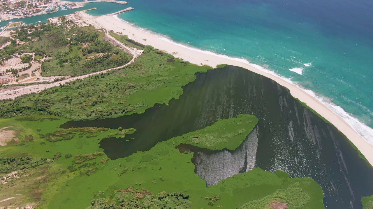Later frame over San Jose del Cabo with more foreground green shoreline visible below the lagoon, the beach running diagonally behind it, and no visible people.