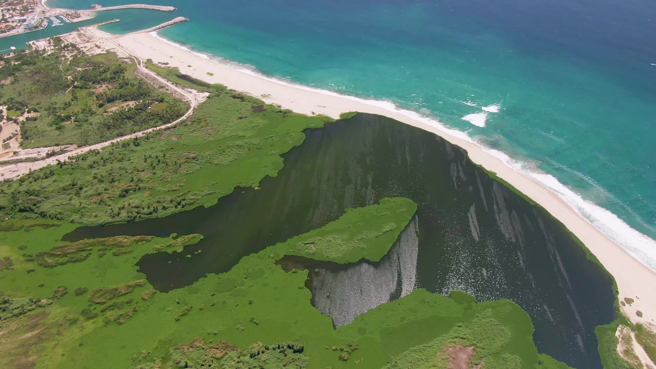 Aerial view in San Jose del Cabo, Baja California Sur, with green cliffs around dark water, a white sandy beach, turquoise ocean, and no visible people.