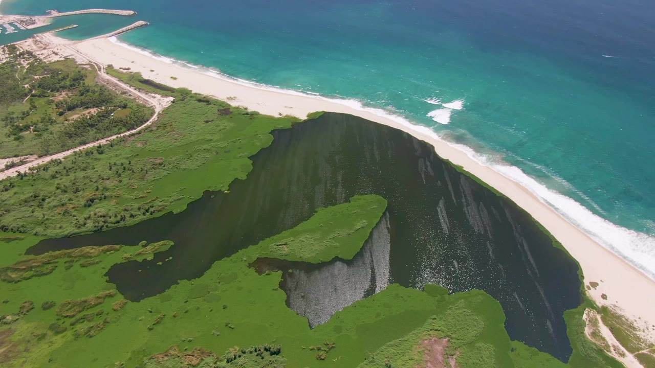 High aerial frame in San Jose del Cabo with turquoise sea, a breaking white surf patch, sandy shoreline, green cliffside vegetation, and no visible people.