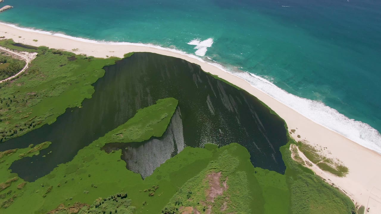 Third aerial frame over San Jose del Cabo shoreline showing a dark water channel behind the sand, bright turquoise sea, foam lines, green dunes, and no visible people.