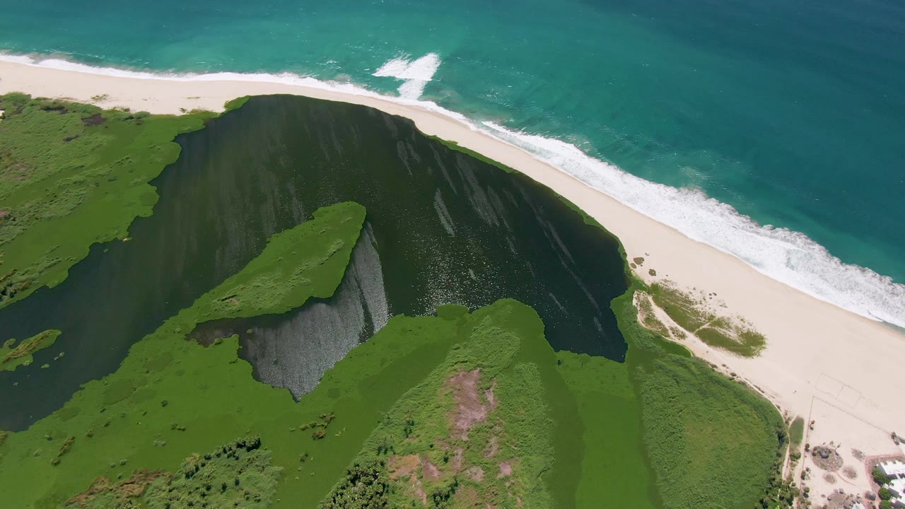 Aerial view in San Jose Del Cabo, Mexico, showing a green cliff above pale sand and turquoise ocean foam, with no visible people.