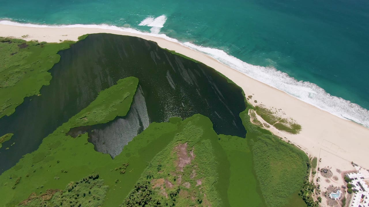 Aerial view of the San Jose Del Cabo coast with a dark green cliff slope, a bright sand strip, turquoise water, and no visible people.