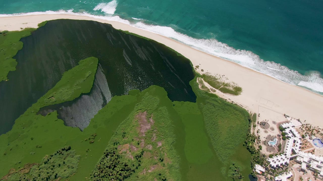 Second aerial frame in San Jose del Cabo with the shadowed green hillside centered, surf breaking along the upper beach, and no visible people.