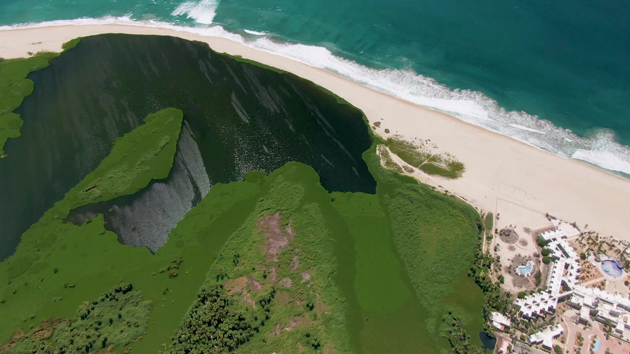 Third aerial frame over San Jose del Cabo showing the steep green ridge, darker shadow mass on the slope, bright sand beach, turquoise ocean, resort buildings, and no visible people.