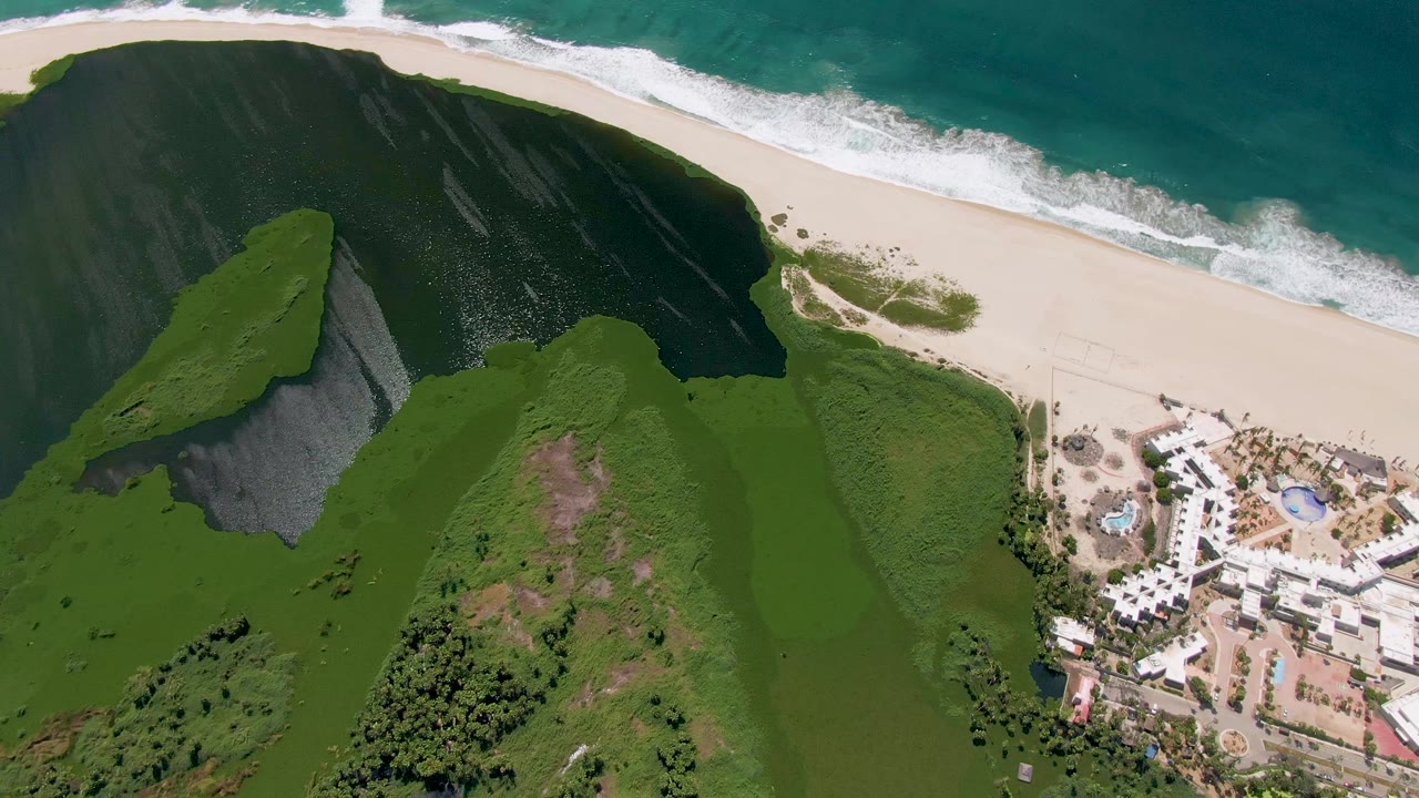 Aerial frame over San Jose Del Cabo with a green hillside and dark rocky cliff beside a pale beach, ocean surf at the top, resort buildings at right, and no visible people.