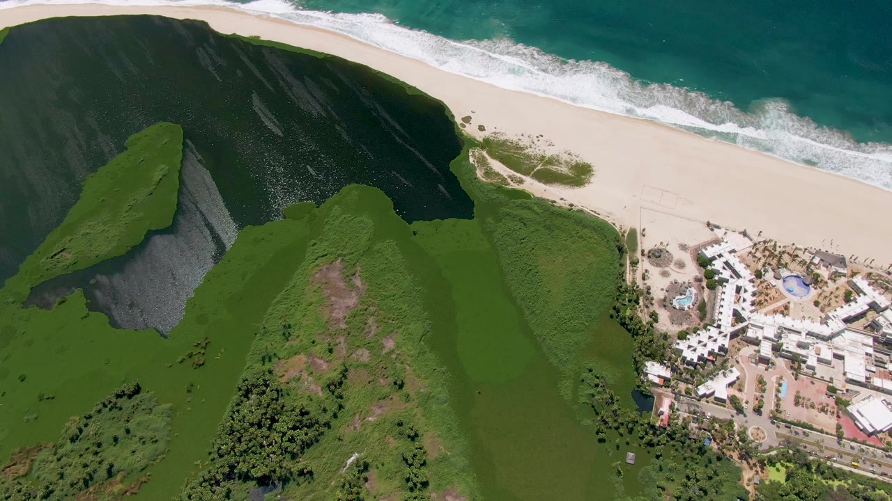 San Jose Del Cabo aerial frame with the dark cliff mass at left, bright green hillside across the center, beach and surf along the top, and the resort complex partly cropped at right, no visible people.
