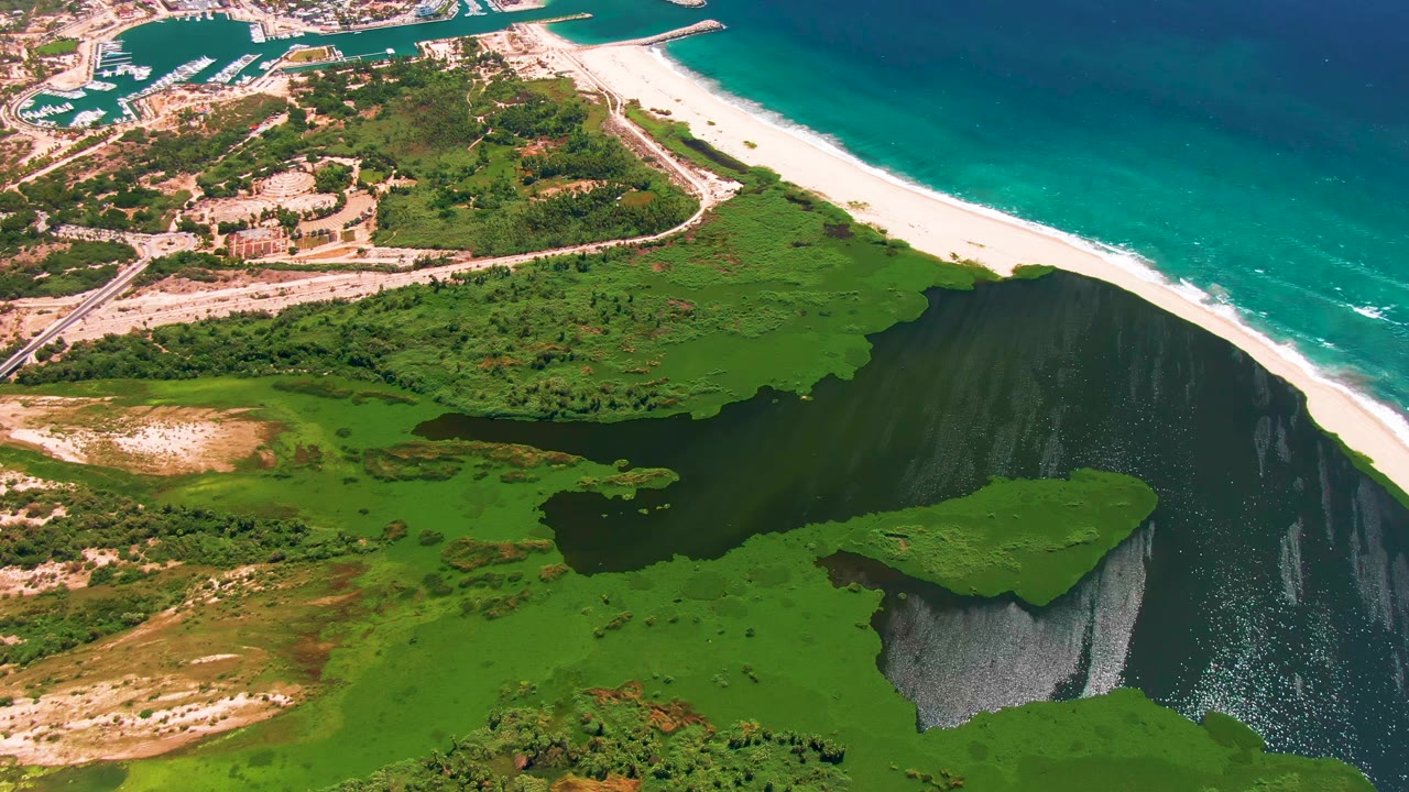 Opening aerial frame over San Jose del Cabo shows dark lagoon water and green vegetation below a long white beach and turquoise ocean, with roads and buildings in the distance and no visible people.