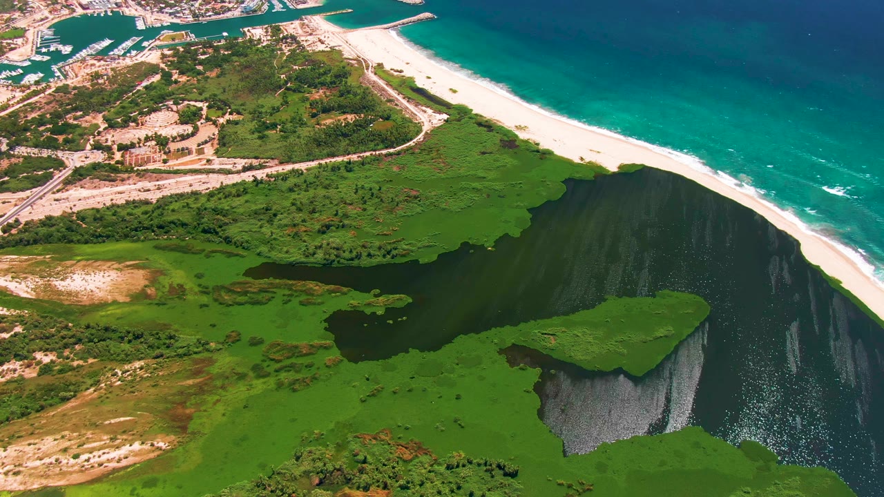 Elevated coastal view with green lagoon edge, sandy beach, ocean surf, and developed shoreline.