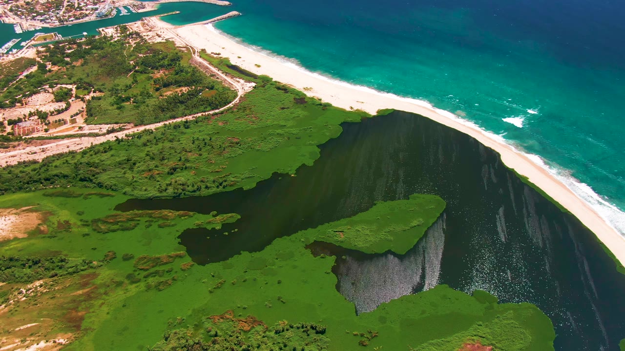 Opening aerial frame over San Jose del Cabo, Mexico, showing white sand beach, turquoise ocean, dark lagoon water, green cliffs, coastal vegetation, and no visible people.