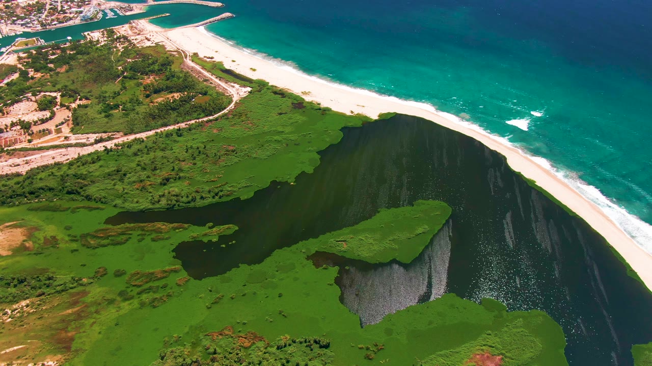 Aerial view in San Jose del Cabo, Mexico, with the lagoon centered below the beach, a green island-like landform, turquoise water offshore, and no visible people.
