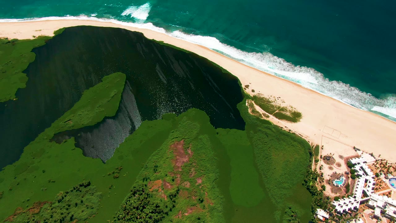 Aerial view of green coastal cliff, sandy beach, ocean surf, and resort edge.