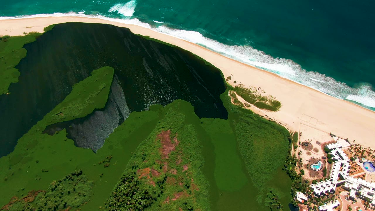 Aerial frame in San Jose del Cabo with the dark green cliff centered closer to the beach, surf breaking along the upper shoreline, resort pools at lower right, and no visible people.