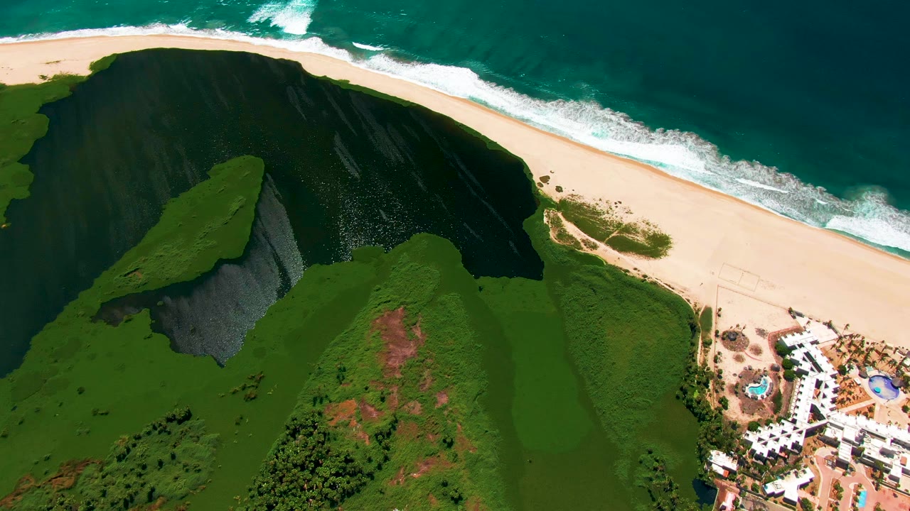 Aerial frame over San Jose del Cabo with surf rolling across the top, a broad sandy beach below it, a green cliff and vegetation in the middle, resort structures at right, and no visible people.