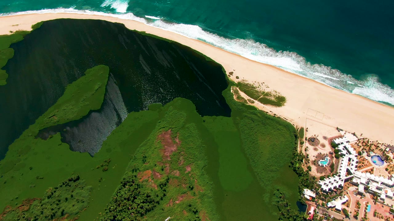 Aerial frame in San Jose del Cabo looking down across the green cliff in the foreground, pale beach and white surf at the top, resort buildings at lower right, and no visible people.