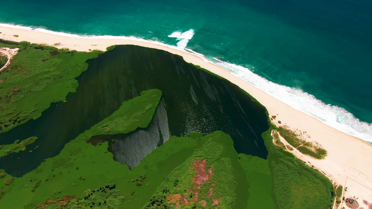 Opening aerial frame over San Jose Del Cabo shows a green cliff dropping toward a pale sand beach and turquoise ocean with white surf, with no visible people.
