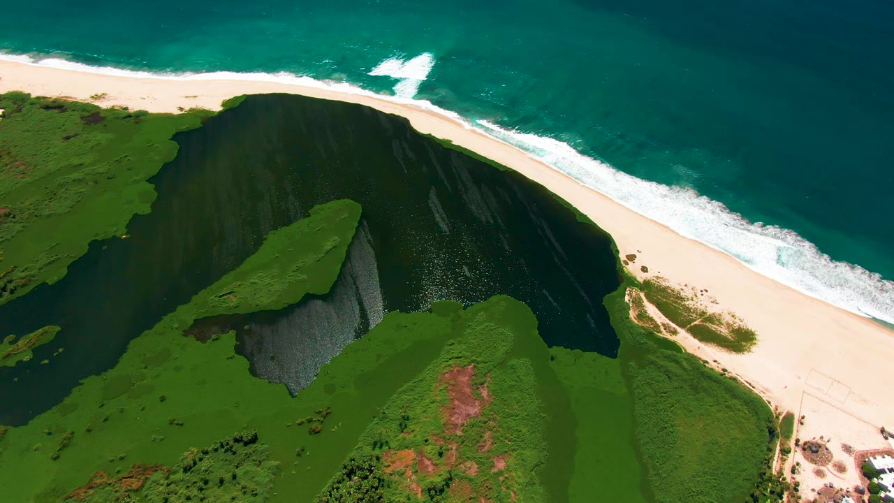 At 0:02, the overhead San Jose Del Cabo view shows the beach curving beside the turquoise water, with the green headland and dark cliff face filling the lower frame, with no visible people.