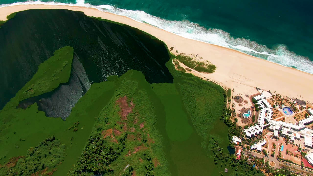 Aerial frame from San Jose del Cabo, Baja California Sur, showing green hills below a sandy beach and blue-green surf, with white resort buildings and pools on the right and no visible people.