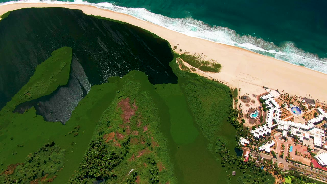 Aerial frame from San Jose del Cabo with the beach and surf running across the top, green hills in the lower frame, and resort pools and white buildings still prominent at the right, with no visible people.