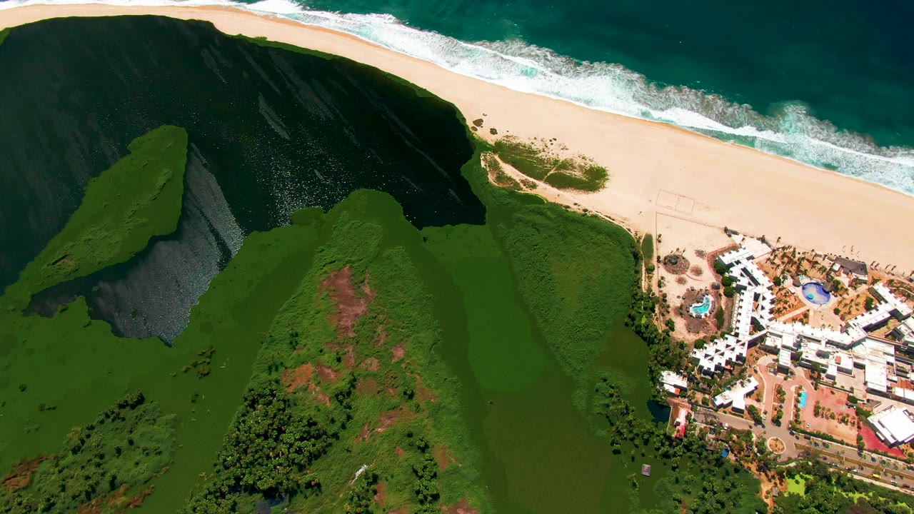 Aerial frame from San Jose del Cabo showing a diagonal surf line above pale sand, a broad green hillside below, and the resort complex beginning to sit closer to the right edge, with no visible people.