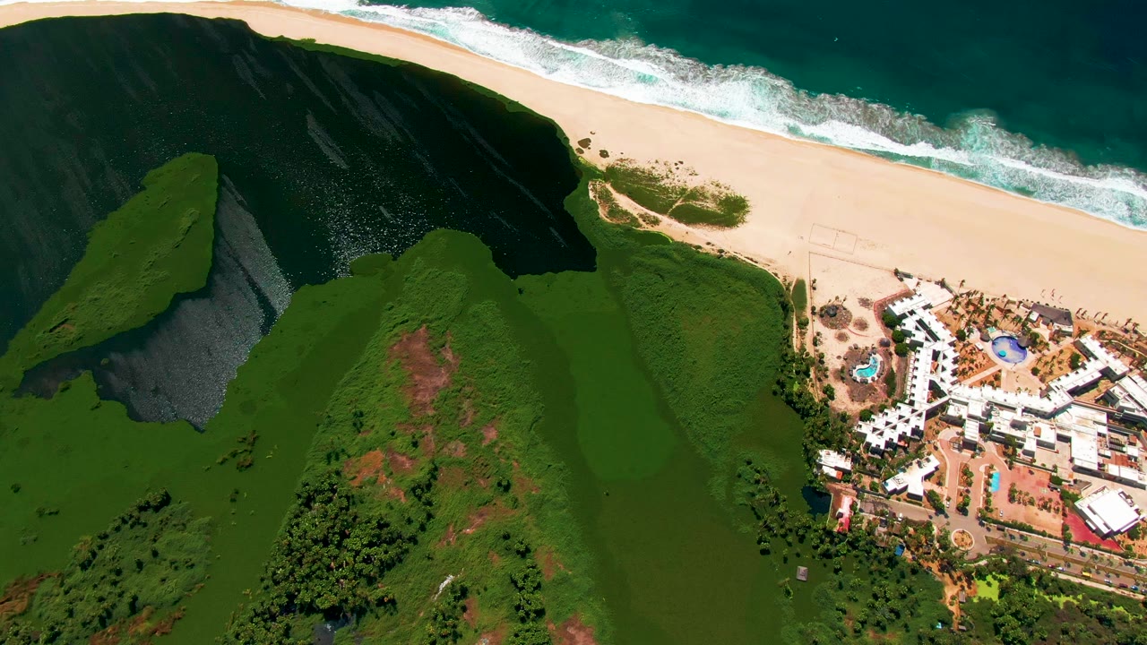 Aerial coastal landscape with green hills, beach, ocean surf, and resort buildings at the right edge.