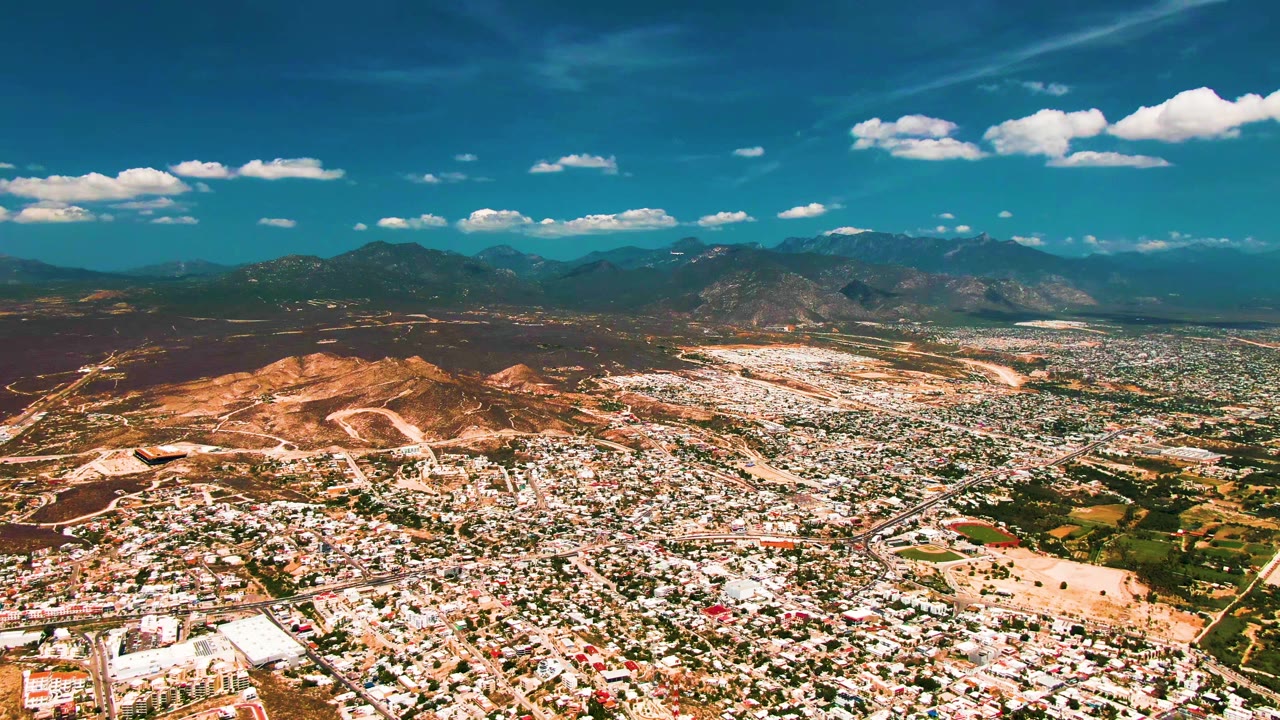 Wide aerial view over San Jose del Cabo with dense city blocks, dry hills, distant mountains, blue sky, scattered white clouds, and no visible people.