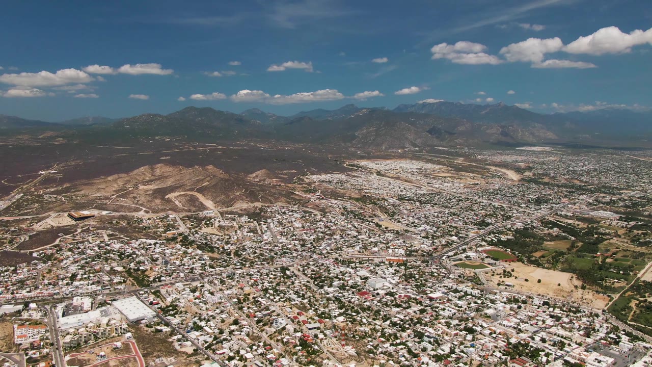 Opening aerial view over San Jose del Cabo, Baja California Sur, showing dense white-roofed buildings, roads, dry hills, distant mountains, blue sky, and no visible people.