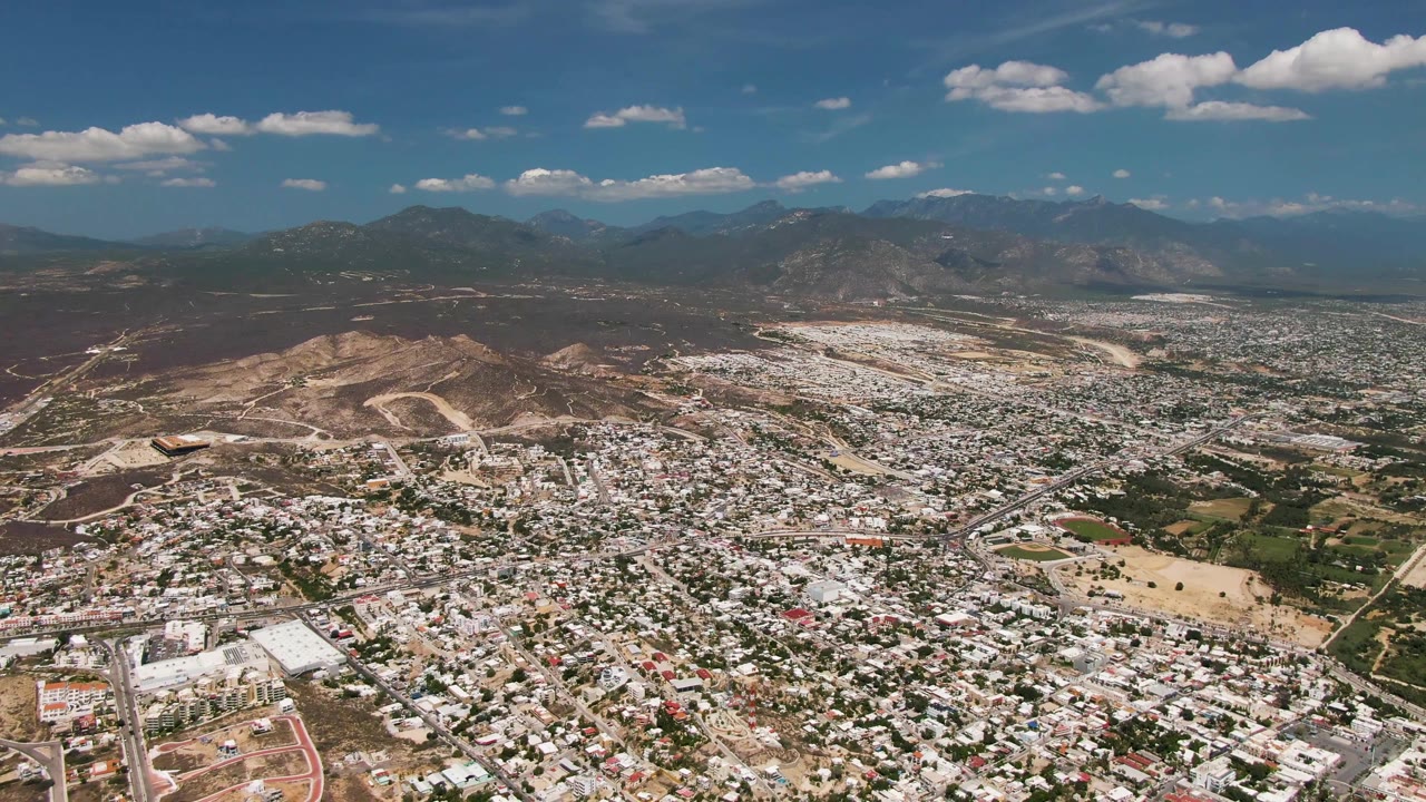 Mid-clip aerial view of San Jose del Cabo, Baja California Sur, with compact white-roofed neighborhoods, a green field near the lower right, dry hills, far mountains, and no visible people.