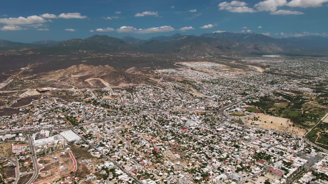 Opening aerial frame over San Jose del Cabo, Mexico, showing dense pale rooftops, roads, dry hills, distant mountains, scattered clouds, and no visible people.