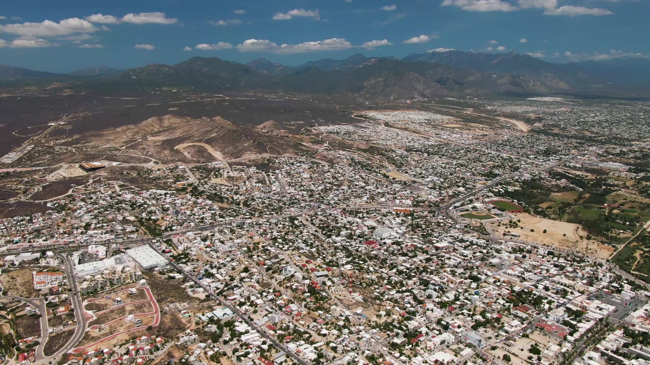 Mid-clip aerial view of San Jose del Cabo showing packed buildings, winding roads near the lower frame, dry terrain around the city, distant mountains, and no visible people.