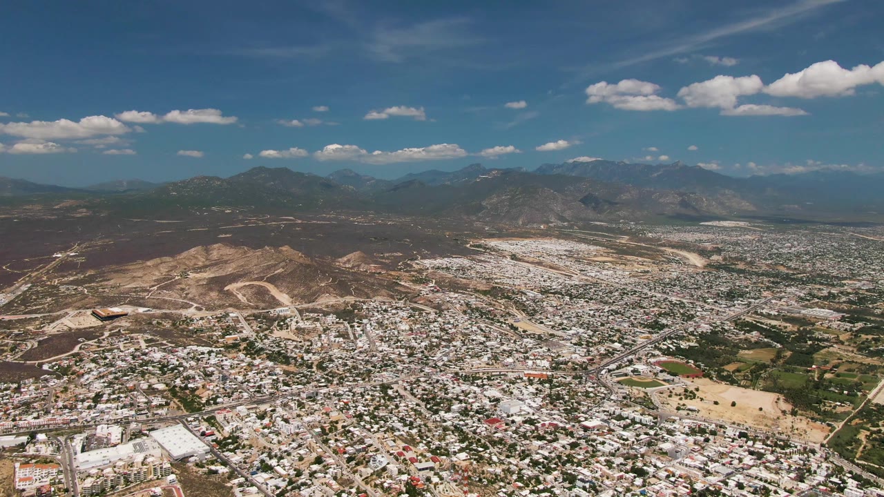 One second into the San Jose del Cabo aerial view, the frame holds the urban foreground and brown hillside midground beneath a mountain horizon and drifting white clouds, with no visible people.