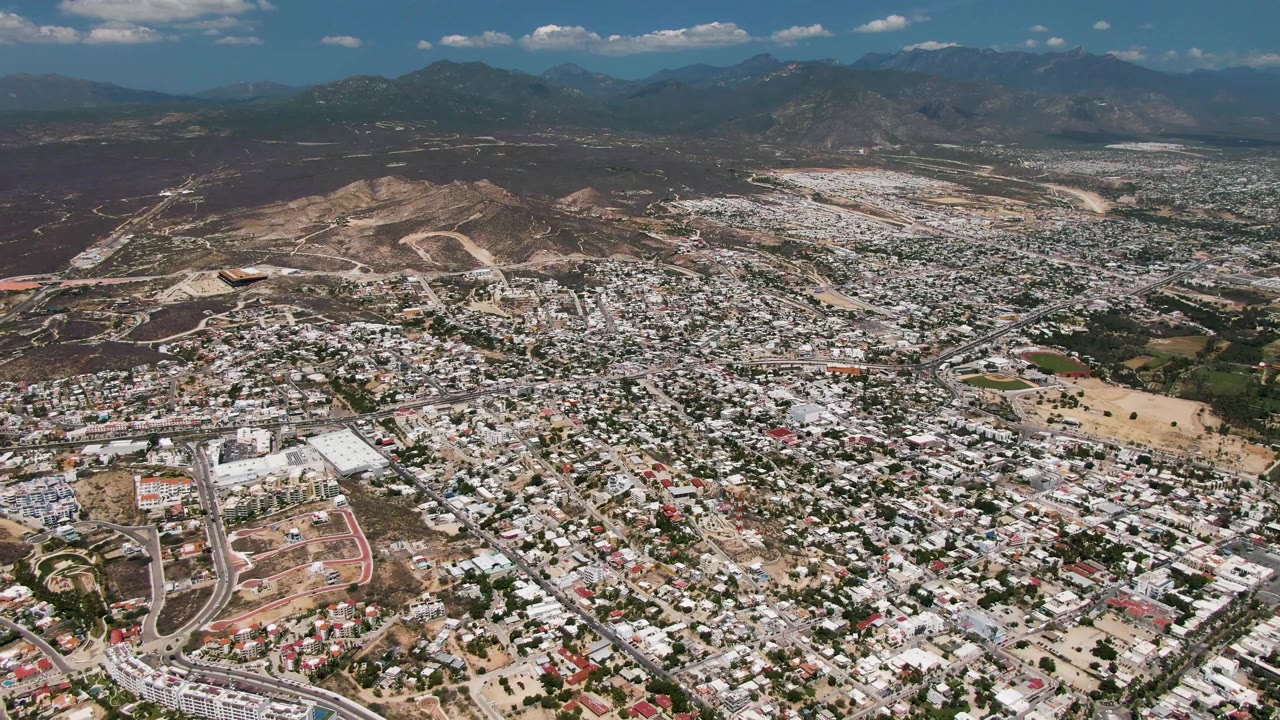 Wide aerial view over San Jose Del Cabo with dense neighborhoods, curving roads, dry hills, distant mountains, scattered clouds, and no visible people.