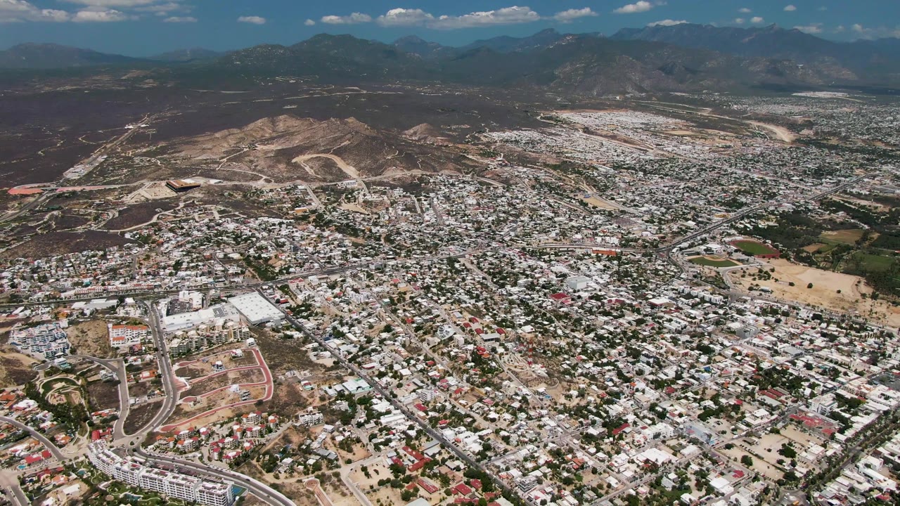 Aerial frame over San Jose Del Cabo showing white and tan rooftops, road corridors, arid hillside slopes, distant mountains, patchy clouds, and no visible people.