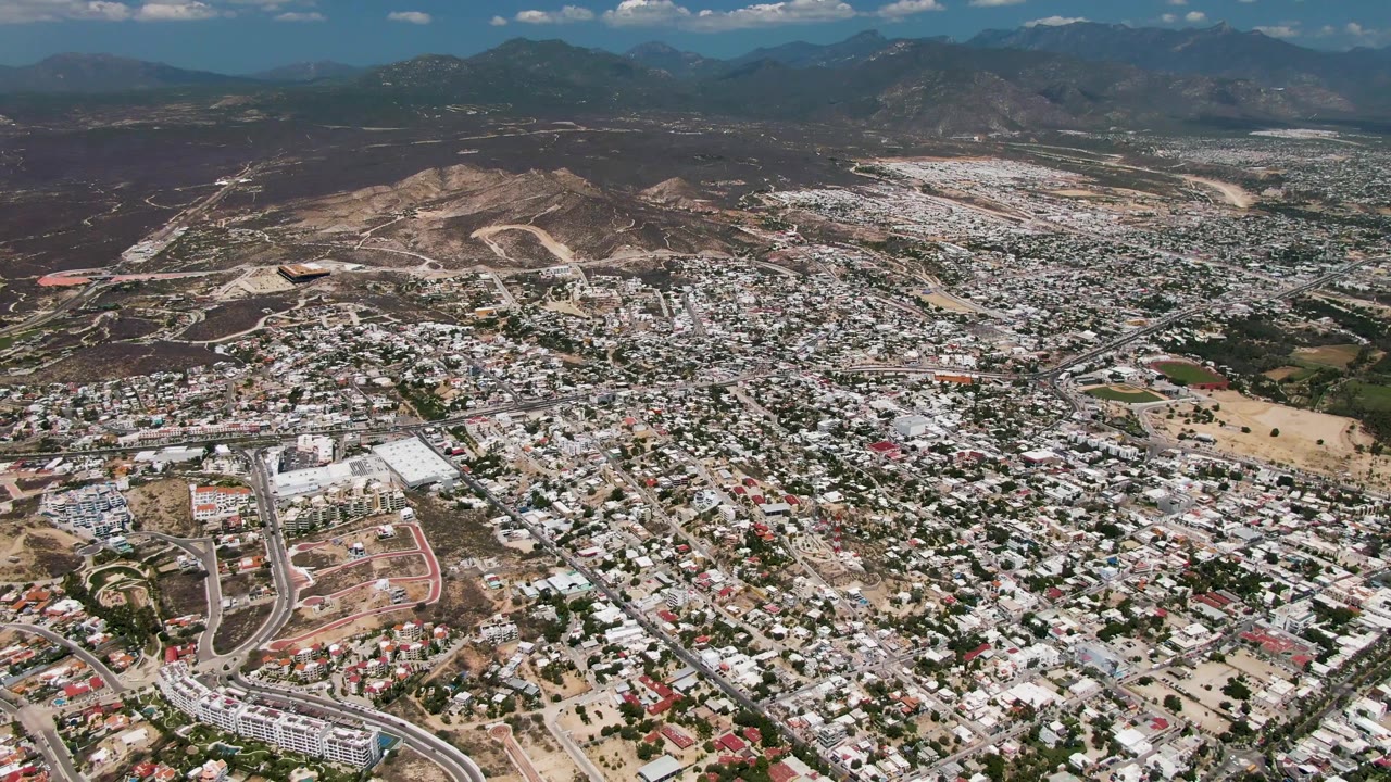 Wide overhead view of San Jose Del Cabo with compact buildings, a pale road interchange in the lower frame, dry brown hills, distant blue mountains, and no visible people.