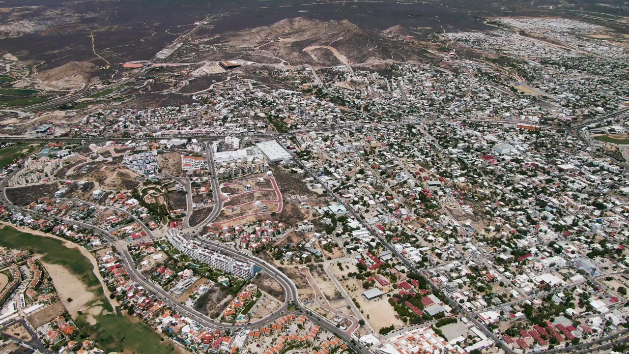Early aerial frame over San Jose Del Cabo residential blocks, curved roads, green spaces, open dirt lots, and dry hills in the background, with no visible people.