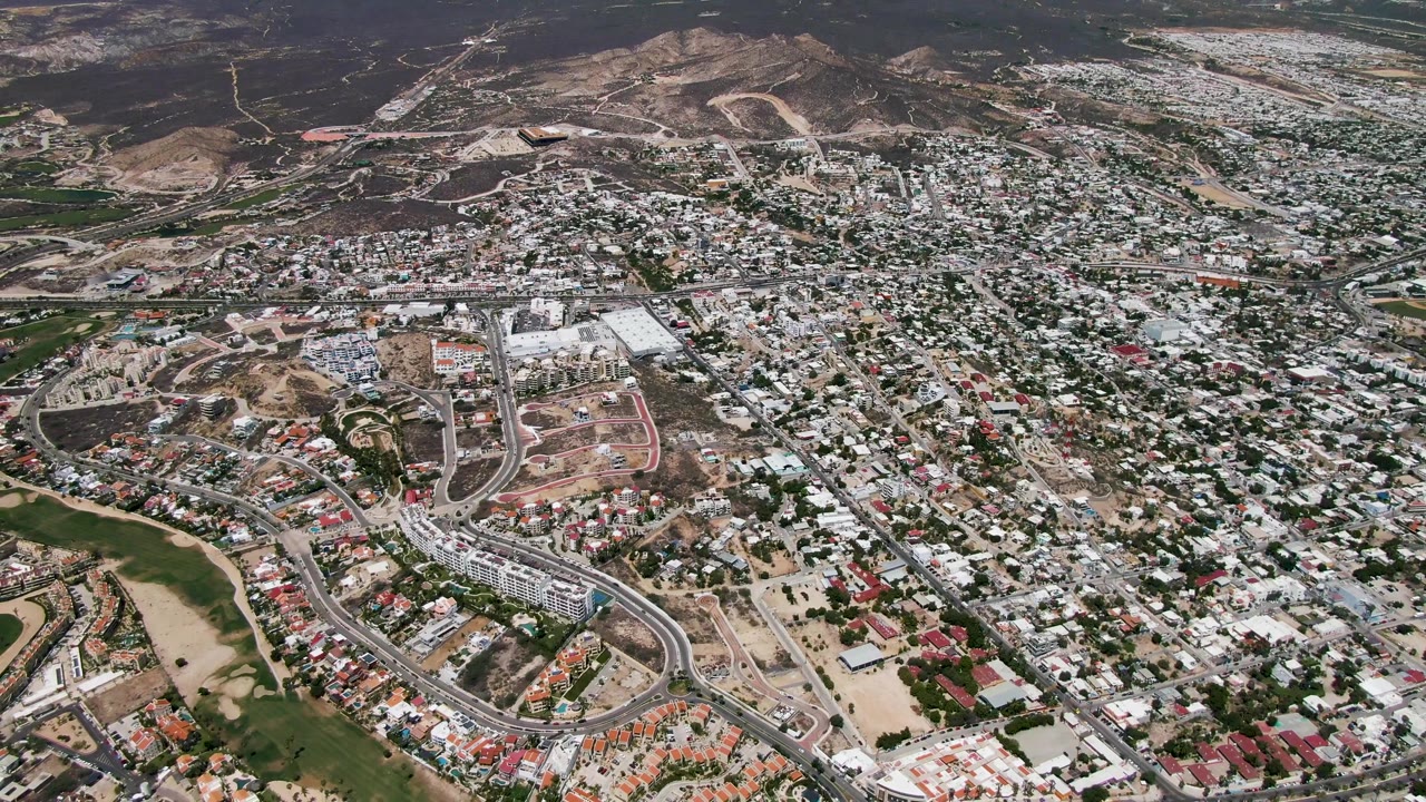 One second into the San Jose Del Cabo aerial, the frame centers on dense rooftops, winding streets, green patches, and tan construction lots, with no visible people.